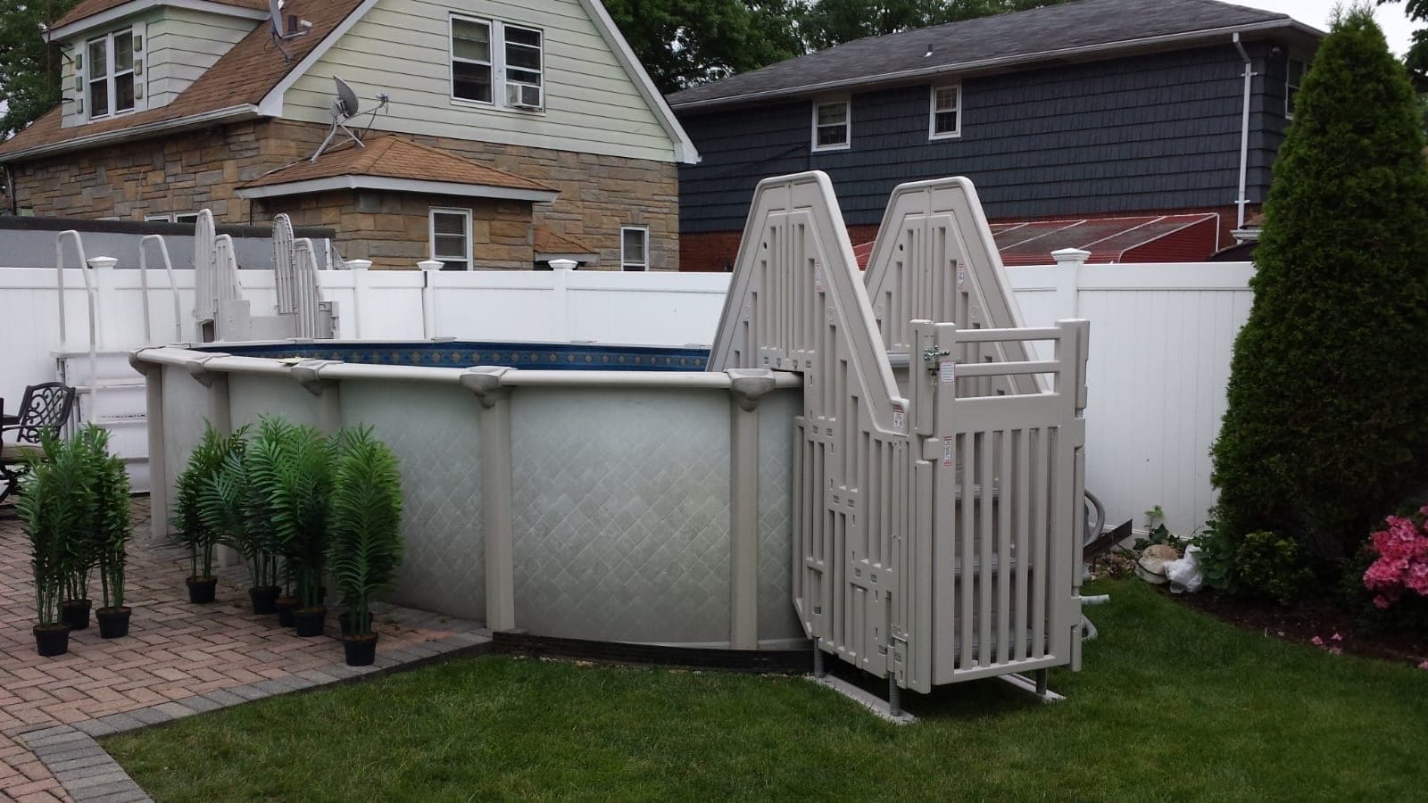 Taupe entry system with a latching gate on the outside of an oval pool, its steps coming down to the lawn.