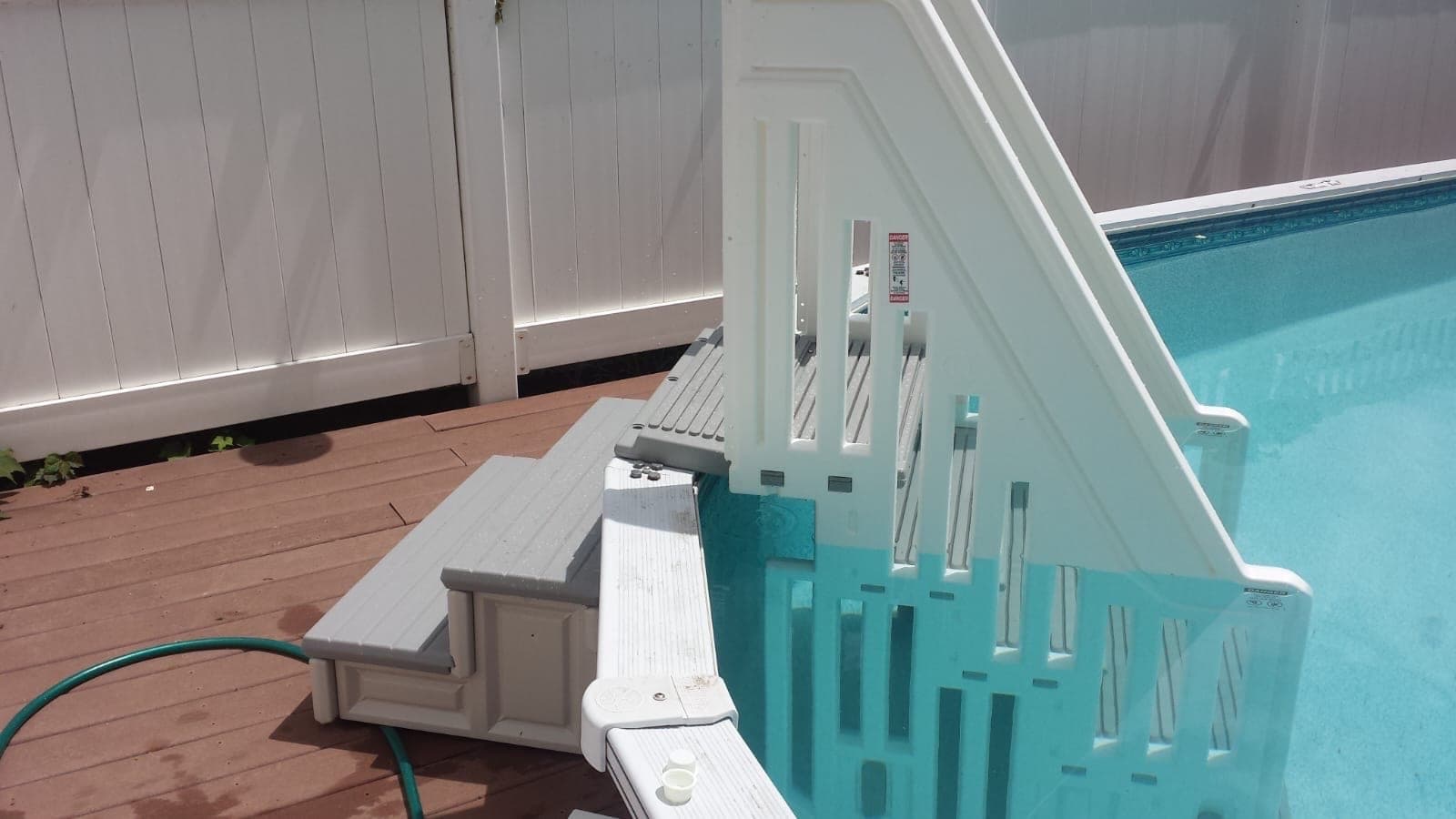 Grey-tread entry steps at a deck edge, the handrail carrying its warning label, water just past the bottom step.