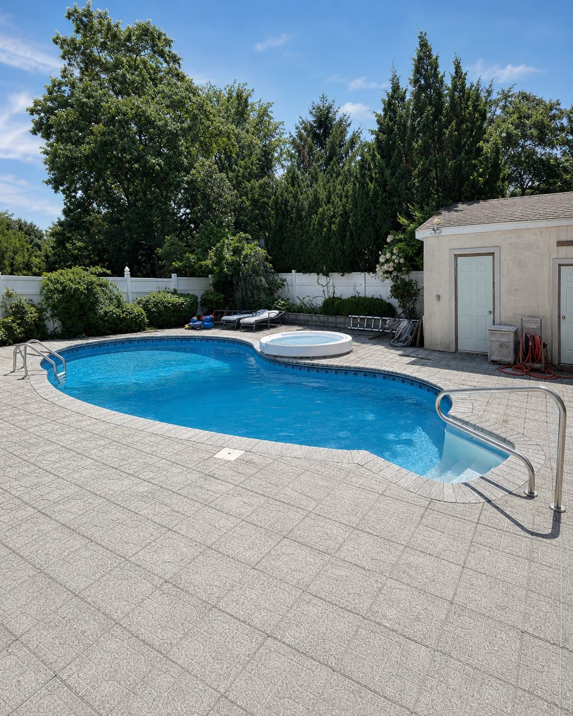 In-ground kidney pool with attached round spa, stone paver deck, white privacy fence, and adjacent poolhouse.
